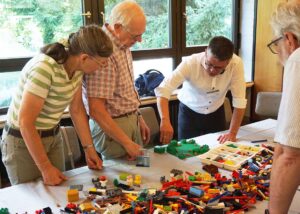 Drei Personen arbeiten gemeinsam an einem Tisch mit bunten Legosteinen und gestalten eine kreative Szene.