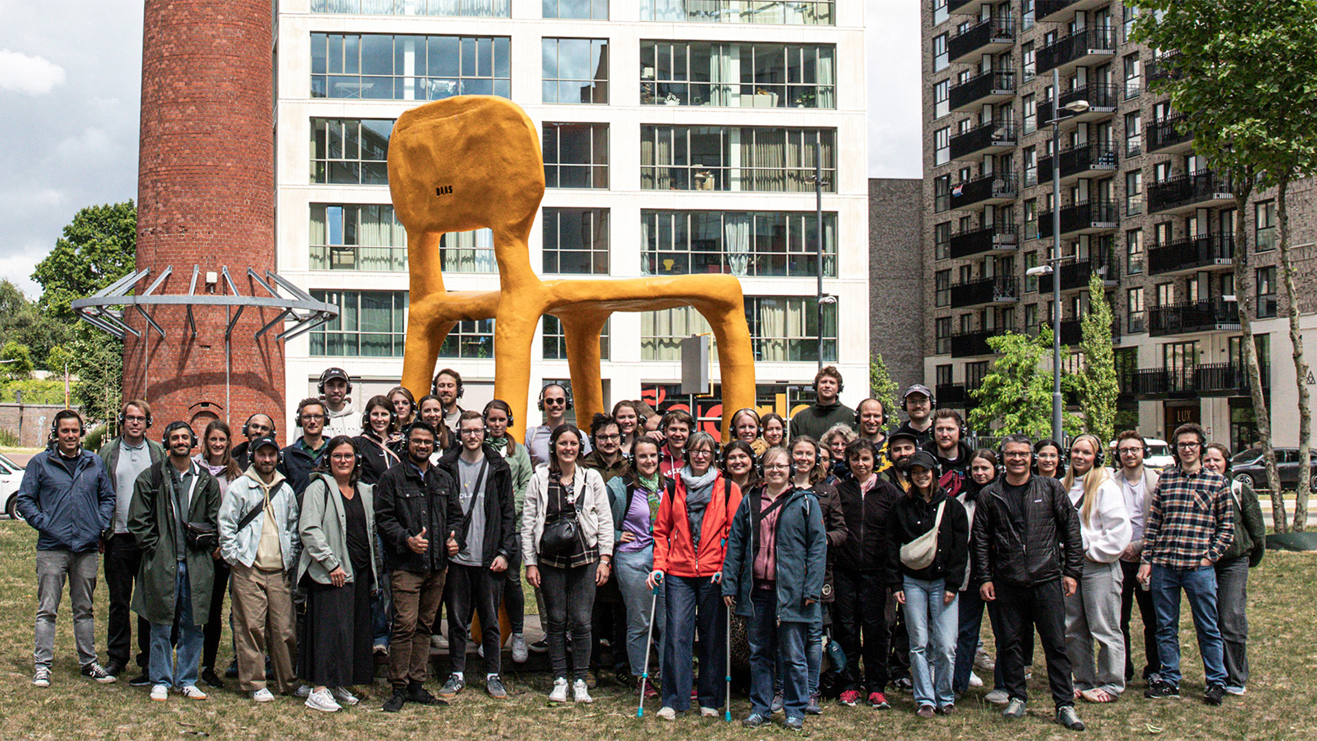 Eine gro&szlig;e Gruppe von Menschen posiert vor einer &uuml;bergro&szlig;en, gelben Stuhlskulptur und modernen Geb&auml;uden.