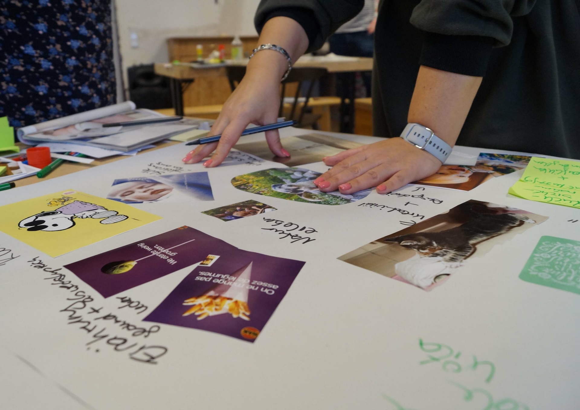 Eine Hand legt Fotos und Zeichnungen auf einen Tisch, w&auml;hrend Notizen und Bastelmaterialien im Hintergrund sichtbar sind.