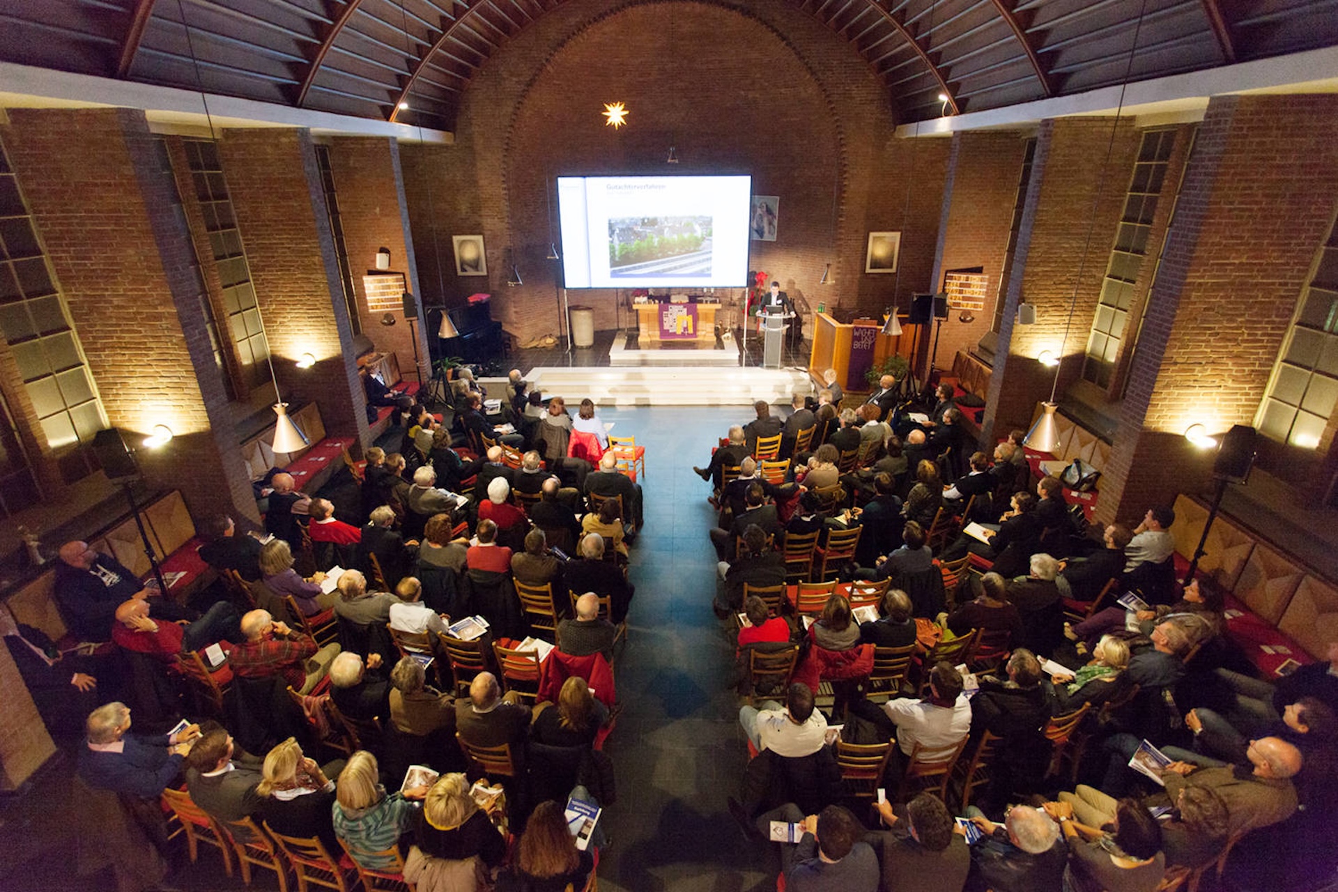 Eine gro&szlig;e Gruppe von Menschen sitzt in einem Saal, w&auml;hrend ein Vortrag auf einer Leinwand gezeigt wird.