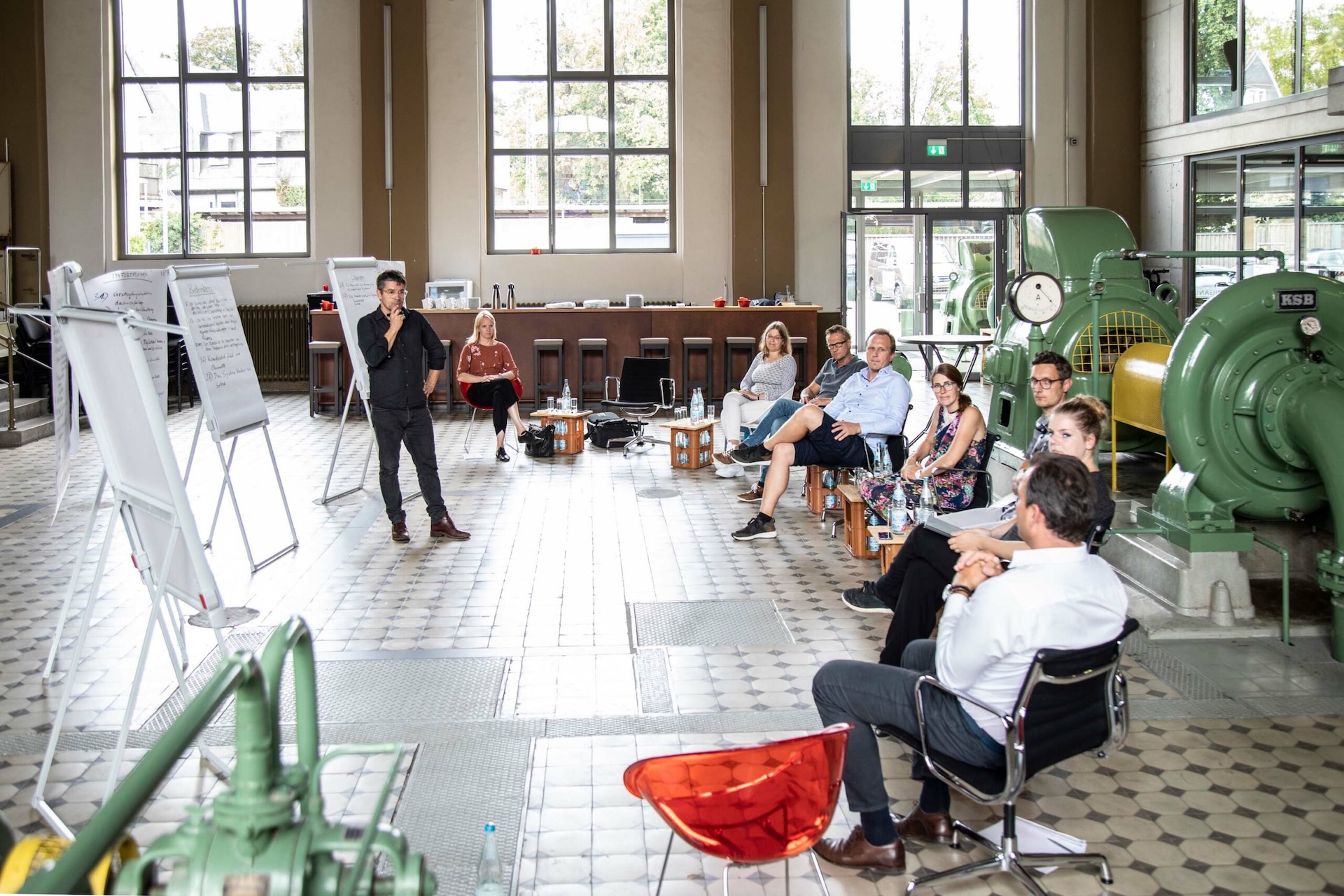 Eine Gruppe von Menschen sitzt in einem modernen Raum, w&auml;hrend ein Mann eine Pr&auml;sentation vor Flipcharts h&auml;lt.