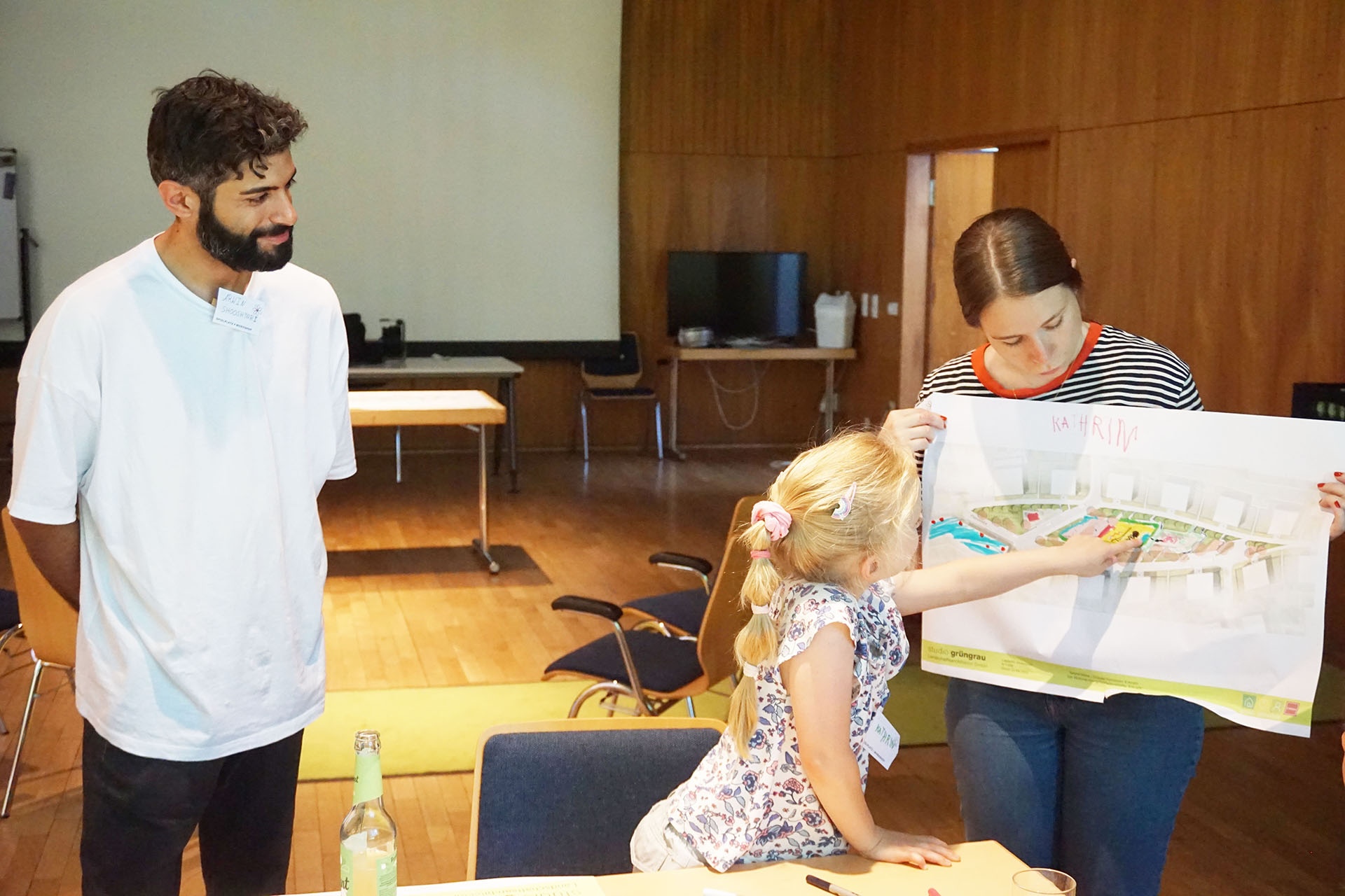 Ein Mann und eine Frau zeigen einem M&auml;dchen einen Plan in einem Konferenzraum. Holzverkleidung und Tische im Hintergrund.
