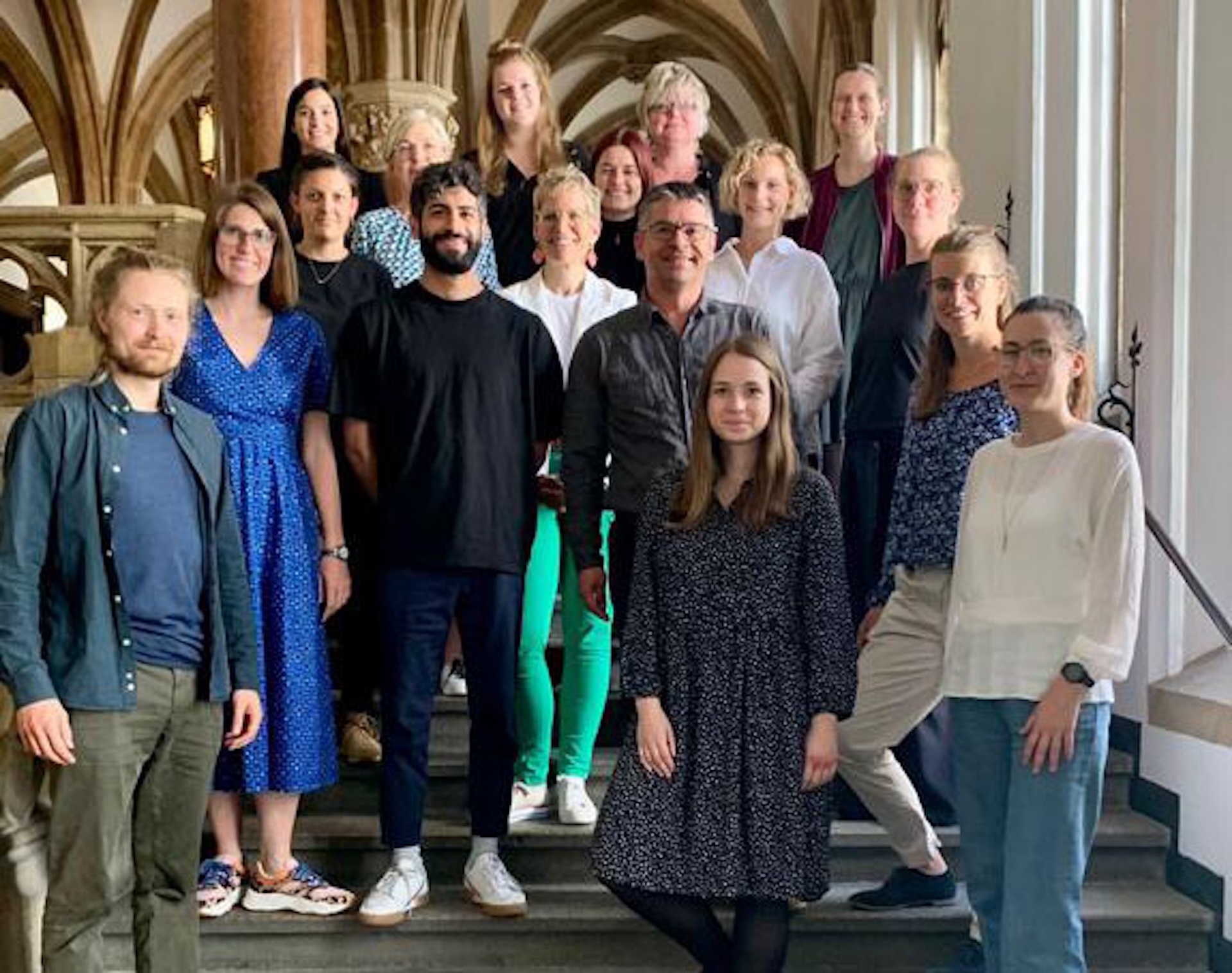 Gruppe von 18 Personen, die auf einer Treppe posieren, in einem historischen Geb&auml;ude mit S&auml;ulen und gro&szlig;en Fenstern.