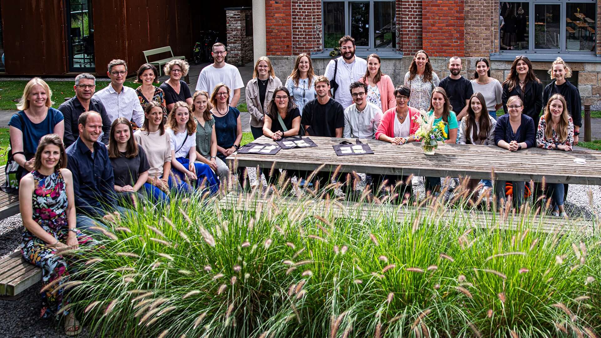 Eine Gruppe von 30 Personen posiert l&auml;chelnd an einem Holztisch in einem Garten vor einem historischen Geb&auml;ude.
