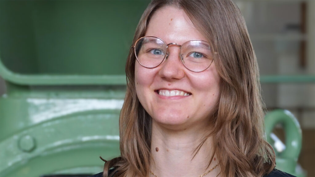 Eine lächelnde Frau mit Brille, braunen Haaren und einem grünen Hintergrund.